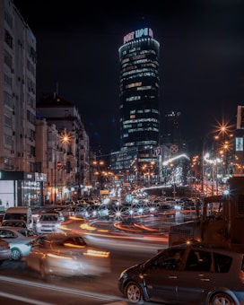 A bustling nighttime cityscape featuring a tall illuminated skyscraper with 'PORT LIFE' signage at the top. The scene is vibrant with heavy traffic, showcasing numerous car light trails that emphasize motion and activity. Surrounding buildings line the street, and street lamps cast a warm glow over the area.
