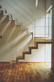 A modern, minimalist staircase with wooden steps and white metal railings casts geometric shadows on the adjacent walls. Natural light streams in from a nearby large window, illuminating the hardwood floor.
