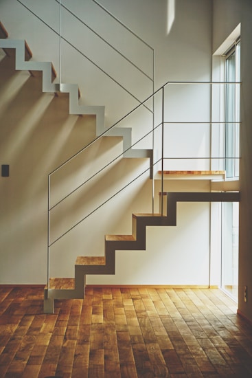 A modern, minimalist staircase with wooden steps and white metal railings casts geometric shadows on the adjacent walls. Natural light streams in from a nearby large window, illuminating the hardwood floor.