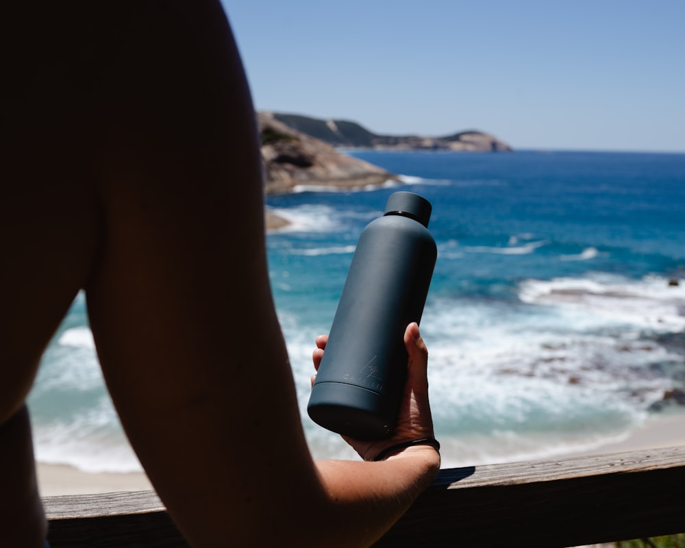 person holding black bottle on brown wooden table near sea during daytime