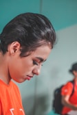 A person with short, dark hair is captured in profile, gazing downwards with a calm and focused expression. They are wearing a bright orange shirt. In the background, there is a slightly blurred figure wearing a similar orange shirt and a backpack, suggesting a group or team setting.