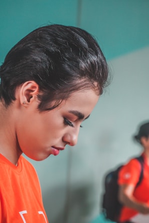 A person with short, dark hair is captured in profile, gazing downwards with a calm and focused expression. They are wearing a bright orange shirt. In the background, there is a slightly blurred figure wearing a similar orange shirt and a backpack, suggesting a group or team setting.