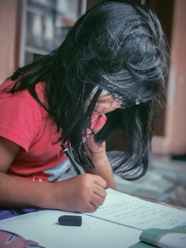 A young girl with long dark hair, wearing glasses and a red shirt, is focused on writing in a notebook. An eraser is visible nearby on the notebook.
