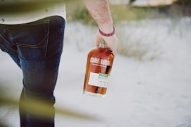 A person holding a bottle of whiskey outdoors, with a sandy ground and some greenery in the background. The person is wearing jeans and a white shirt, and the whiskey bottle has a prominent label.