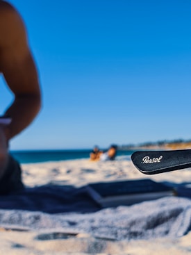 A sandy beach scene with a clear blue sky in the background. A person is partially visible on the left side, sitting on a towel. In the foreground, a pair of sunglasses with the brand name 'Persol' is prominently displayed. Two people can be seen sitting further away near the shoreline.