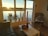 A cozy living room features a large window overlooking a serene lake during sunset. There are several potted plants on the windowsill, a black wicker chair with a patterned cushion, and a coffee table with decorative items. The room has wooden flooring and a modern, minimalist design.