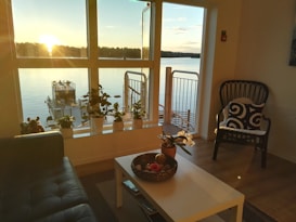 A cozy living room features a large window overlooking a serene lake during sunset. There are several potted plants on the windowsill, a black wicker chair with a patterned cushion, and a coffee table with decorative items. The room has wooden flooring and a modern, minimalist design.