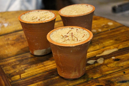 Three ceramic cups filled with a frothy beverage are placed on a wooden table. The drink has visible strands of saffron on top, and the cups are unglazed, exhibiting a rustic and earthy texture.