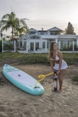 A woman is inflating a stand-up paddleboard on a sandy surface. She is wearing a white swimsuit and standing barefoot next to the board. A large house with glass doors and windows is visible in the background, along with palm trees and shrubs.