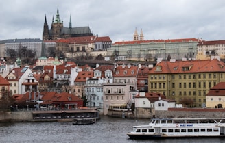 A scenic view of a historic city featuring an ornate castle with tall spires and a mix of Renaissance and Gothic architecture. Below the castle, there are charming houses with red-tiled roofs and a large river in the foreground with a tourist boat labeled 'JAZZ BOAT.' The sky is overcast, adding a moody atmosphere to the scene.
