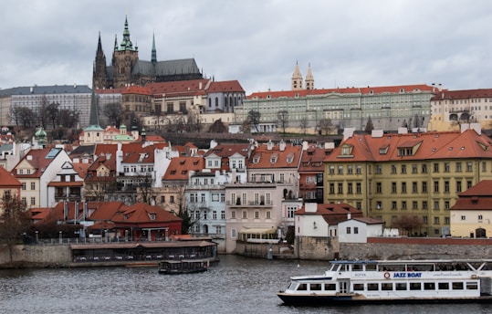 A scenic view of a historic city featuring an ornate castle with tall spires and a mix of Renaissance and Gothic architecture. Below the castle, there are charming houses with red-tiled roofs and a large river in the foreground with a tourist boat labeled 'JAZZ BOAT.' The sky is overcast, adding a moody atmosphere to the scene.