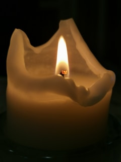 A partially melted candle with a bright flame flickers gently, casting a warm glow. The wax has formed organic, flowing shapes around the top.