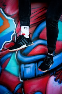 Two legs in black pants are wearing black sneakers and white socks with the number 1343 and cherry graphics. They are positioned against a vibrant, colorful graffiti wall with abstract shapes and lines in shades of red, blue, and teal.