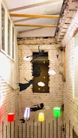 A narrow space with exposed brick walls features a creative installation depicting silhouettes of flying children and objects on the wall. Colorful lamps at different heights add a whimsical element, casting vibrant red, green, and yellow light against the neutral backdrop.