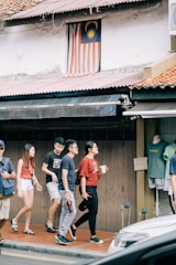 A group of people walks along a sidewalk near a building featuring a Malaysian flag displayed in a window above. They pass a shop with clothing displayed on a mannequin. The scene is urban, with an awning and a parked vehicle partially visible in the foreground.