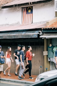A group of people walks along a sidewalk near a building featuring a Malaysian flag displayed in a window above. They pass a shop with clothing displayed on a mannequin. The scene is urban, with an awning and a parked vehicle partially visible in the foreground.