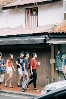 A group of people walks along a sidewalk near a building featuring a Malaysian flag displayed in a window above. They pass a shop with clothing displayed on a mannequin. The scene is urban, with an awning and a parked vehicle partially visible in the foreground.