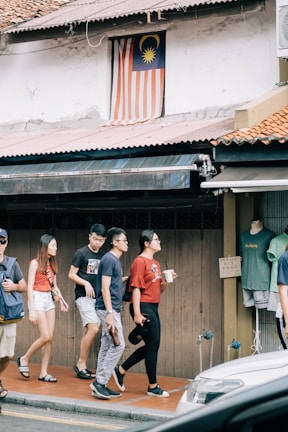 A group of people walks along a sidewalk near a building featuring a Malaysian flag displayed in a window above. They pass a shop with clothing displayed on a mannequin. The scene is urban, with an awning and a parked vehicle partially visible in the foreground.