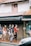 A group of people walks along a sidewalk near a building featuring a Malaysian flag displayed in a window above. They pass a shop with clothing displayed on a mannequin. The scene is urban, with an awning and a parked vehicle partially visible in the foreground.