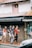 A group of people walks along a sidewalk near a building featuring a Malaysian flag displayed in a window above. They pass a shop with clothing displayed on a mannequin. The scene is urban, with an awning and a parked vehicle partially visible in the foreground.