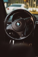 A close-up view of a BMW car steering wheel, showcasing the brand logo in the center and various control buttons on the sides. The background includes a blurred view of another vehicle and elements of the car's interior.