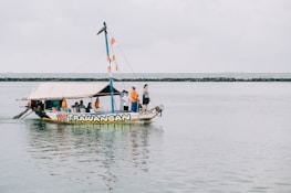 A small, colorful boat with the name 'Gili Trawangan' on it is floating on a calm body of water. Several people are on board, some standing and others seated under a canopy. The boat is decorated with orange and white flags, and the shoreline with a rocky barrier can be seen in the background under a cloudy sky.
