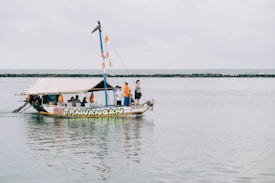 A small, colorful boat with the name 'Gili Trawangan' on it is floating on a calm body of water. Several people are on board, some standing and others seated under a canopy. The boat is decorated with orange and white flags, and the shoreline with a rocky barrier can be seen in the background under a cloudy sky.