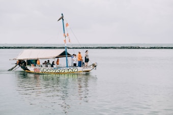 A small, colorful boat with the name 'Gili Trawangan' on it is floating on a calm body of water. Several people are on board, some standing and others seated under a canopy. The boat is decorated with orange and white flags, and the shoreline with a rocky barrier can be seen in the background under a cloudy sky.