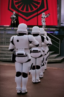 A group of armored soldiers is lined up in formation. They are wearing white suits with black detailing and appear to be marching. In the background, there is a large red banner with a black emblem, set above a stage with other similar soldiers.