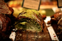 A slice of bread or pastry with a swirl pattern is displayed on a wooden surface. The top is covered with green pistachio crumbs and a drizzle of icing, giving it a lush, textured appearance. The background has a warm, blurred effect, suggesting a bakery setting.