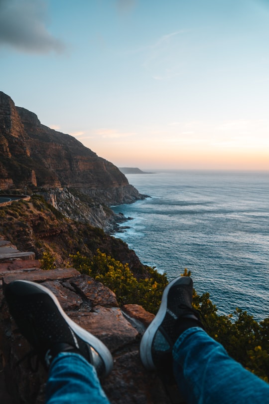 person in blue shirt sitting on rock formation near body of water during daytime in Chapman's Peak Drive South Africa