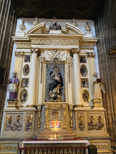 An ornate religious altar with intricate gold and marble details, featuring statues of saints and religious figures. Several candles are lit in front of the altar, contributing to a solemn atmosphere.