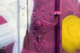 A vintage-style, ornate door handle painted in a deep pink color, attached to a weathered surface with graffiti. The background features a mix of colors including white, red, and a splash of bright yellow.