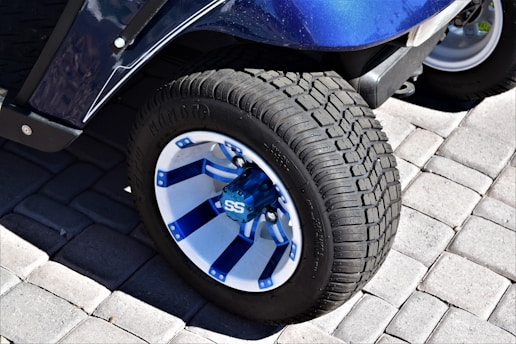 A close-up of a vehicle wheel featuring a blue and white alloy rim with a bold 'SS' logo in the center. The tire has a rough tread pattern, and the wheel is situated on a surface made of interlocking gray pavers.