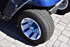 A close-up of a vehicle wheel featuring a blue and white alloy rim with a bold 'SS' logo in the center. The tire has a rough tread pattern, and the wheel is situated on a surface made of interlocking gray pavers.
