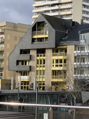 A modern building featuring a distinctive roof with an angular design, contrasted by bright yellow window frames and accents. The facade is a mix of concrete and dark cladding, creating a robust architectural style. Smaller adjacent structures are visible, along with a tree in the foreground and part of another building in the background.