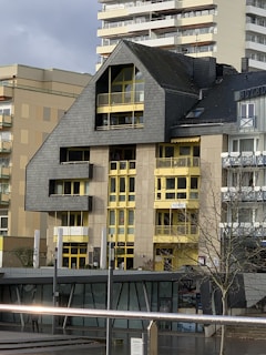 A modern building featuring a distinctive roof with an angular design, contrasted by bright yellow window frames and accents. The facade is a mix of concrete and dark cladding, creating a robust architectural style. Smaller adjacent structures are visible, along with a tree in the foreground and part of another building in the background.