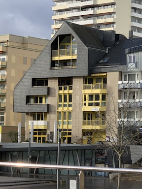 A modern building featuring a distinctive roof with an angular design, contrasted by bright yellow window frames and accents. The facade is a mix of concrete and dark cladding, creating a robust architectural style. Smaller adjacent structures are visible, along with a tree in the foreground and part of another building in the background.