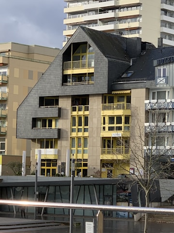 A modern building featuring a distinctive roof with an angular design, contrasted by bright yellow window frames and accents. The facade is a mix of concrete and dark cladding, creating a robust architectural style. Smaller adjacent structures are visible, along with a tree in the foreground and part of another building in the background.