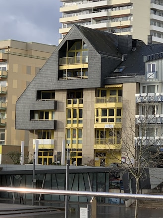 A modern building featuring a distinctive roof with an angular design, contrasted by bright yellow window frames and accents. The facade is a mix of concrete and dark cladding, creating a robust architectural style. Smaller adjacent structures are visible, along with a tree in the foreground and part of another building in the background.