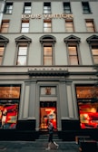 The facade of a Louis Vuitton store featuring large display windows with mannequins dressed in stylish outfits. The building has a classic architectural style with tall windows and decorative elements. A person is walking past the store, which has a logo above the entrance and vibrant colors in the window displays.