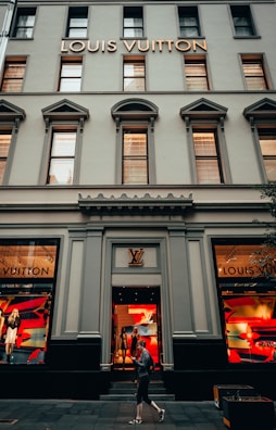 The facade of a Louis Vuitton store featuring large display windows with mannequins dressed in stylish outfits. The building has a classic architectural style with tall windows and decorative elements. A person is walking past the store, which has a logo above the entrance and vibrant colors in the window displays.
