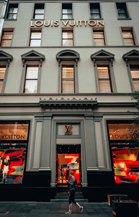 The facade of a Louis Vuitton store featuring large display windows with mannequins dressed in stylish outfits. The building has a classic architectural style with tall windows and decorative elements. A person is walking past the store, which has a logo above the entrance and vibrant colors in the window displays.