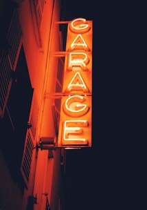 A bright, vertical neon sign spells out the word 'GARAGE' in bold, orange-red letters against a dark background. The sign is mounted on the side of a building, with parts of the structure faintly visible and bathed in warm light.