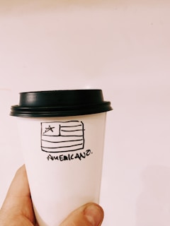 Close-up of a hand holding a coffee mug with a MAGA-style font slogan, set against a backdrop of vintage Americana.