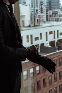 A person in a suit is putting on a glove while standing near a window overlooking a cityscape with tall buildings. The image reflects a professional setting.