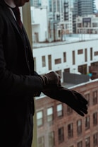A person in a suit is putting on a glove while standing near a window overlooking a cityscape with tall buildings. The image reflects a professional setting.