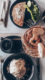 A spread of breakfast dishes includes an omelette with a cucumber slice and lettuce on a black plate, accompanied by a variety of breads and pastries stacked on another plate. A small cup of coffee or tea is placed next to the plates. The setup is on a light wooden table, with silver cutlery arranged neatly on the sides.