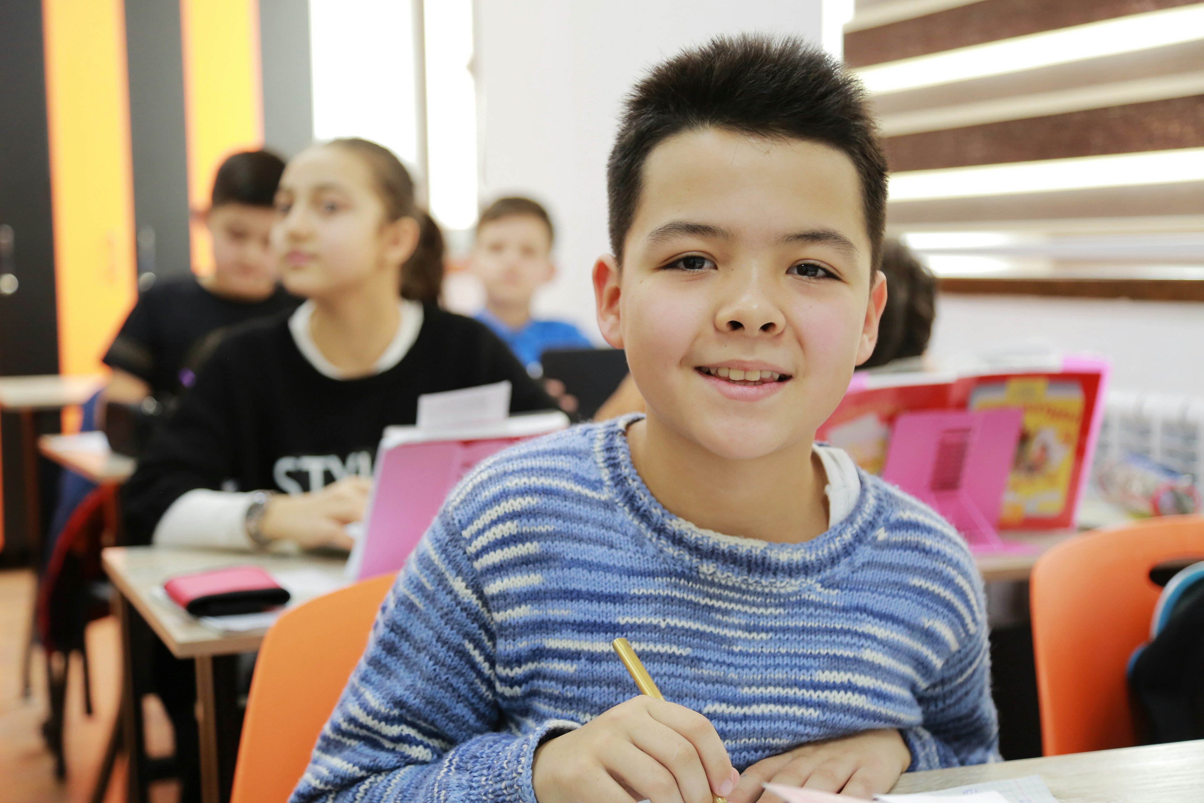 Photo of boy in blue and white striped long sleeve shirt holding pencil while sitting in classroom.