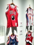A display of several basketball jerseys hanging on a wall, each on wooden hangers. The prominent jersey is red with the word 'BULLS' and a graphic face logo. Other jerseys include a white New York jersey with blue and red accents and a blue jersey with intricate patterns.